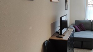 Un salon moderne avec un canapé, un bureau avec un ordinateur et une chaise. Des tableaux encadrés ornent le mur, et une fenêtre est agrémentée de rideaux.