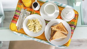 Bandeja de desayuno con huevos revueltos, tostadas, una taza de café y un vaso de jugo de naranja sobre un mantel colorido.