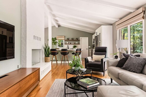 Un salon moderne avec un canapé, une table basse et un téléviseur. On aperçoit un coin repas avec des chaises et, au fond, une cuisine.