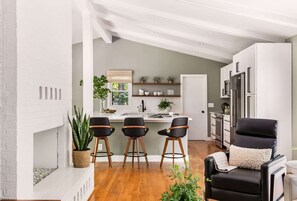 Une cuisine moderne avec des meubles blancs, un îlot central et des tabourets de bar. Le salon est doté d’un canapé en cuir noir agrémenté d’un coussin décoratif.