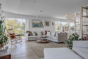 Un salon spacieux avec un canapé blanc, un fauteuil à bascule en bois et une table basse. Une grande fenêtre offre une vue sur les arbres, et un escalier en colimaçon se trouve dans un coin.