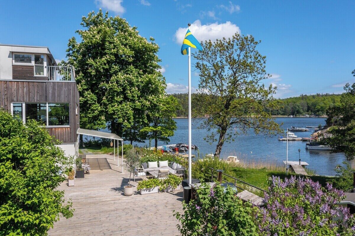 Una casa moderna con una terraza de madera que da a un lago, con barcos amarrados y una bandera.