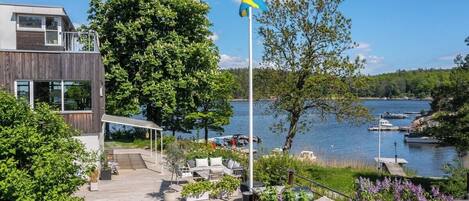 Una casa moderna con una terraza de madera que da a un lago, con barcos amarrados y una bandera.