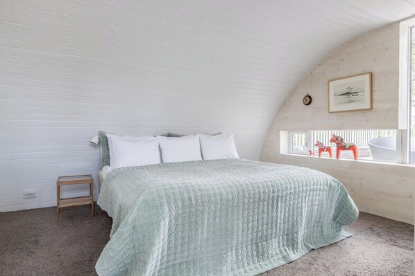A bedroom with a bed, a small wooden stool, a window with a view of a boat, and a framed picture on the wall.