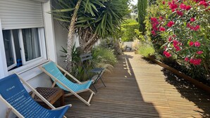 Une terrasse en bois avec deux chaises longues bleues et une petite table, entourée d’une végétation luxuriante et de fleurs aux couleurs vives.