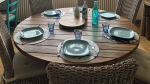 Une table de salle à manger ronde en bois, avec des assiettes, des verres et des bouteilles, placée devant une fenêtre donnant sur un paysage verdoyant et des bâtiments.