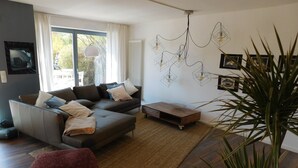 A modern living room with a sofa, ottoman, and a coffee table. There is a large window with curtains, a radiator, and a wall-mounted light fixture. - Ferienbungalow Iwele (Marktbreit)