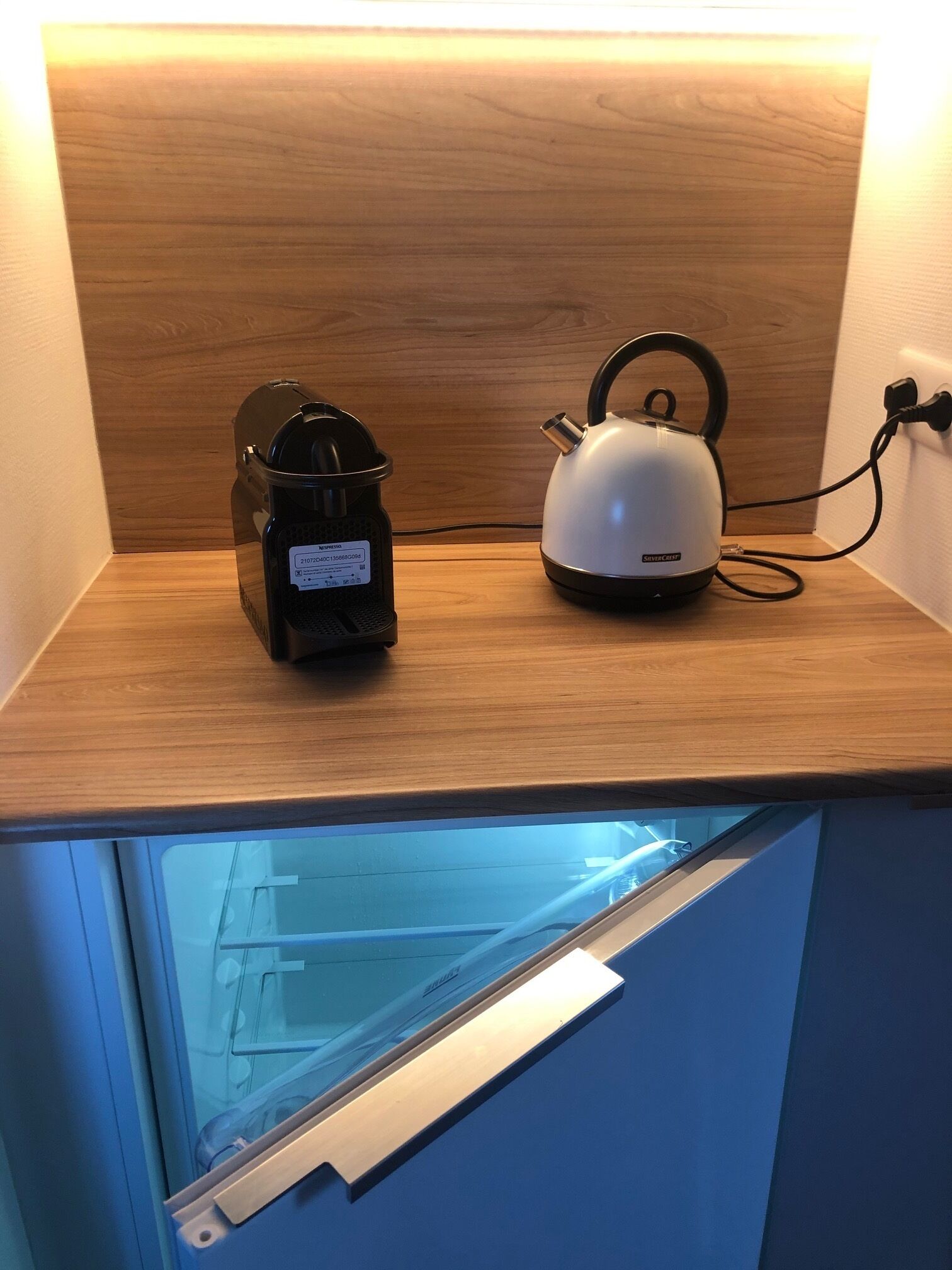 Eine Küchenzeile mit Kaffeemaschine und Wasserkocher auf einer Holzausziehtafel, ein Fenster darunter und eine wandmontierte Steckdose.