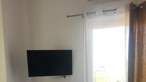 A room with a flat-screen TV mounted on the wall, a small black cabinet, a white curtain, and a view of a balcony with a railing. - Appartement Indépendant - Wifi - Parking- Petit Déjeuner (Ittenheim)