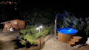 Un jacuzzi rodeado por una terraza de madera, una cabaña pequeña de madera y un sendero de piedra.