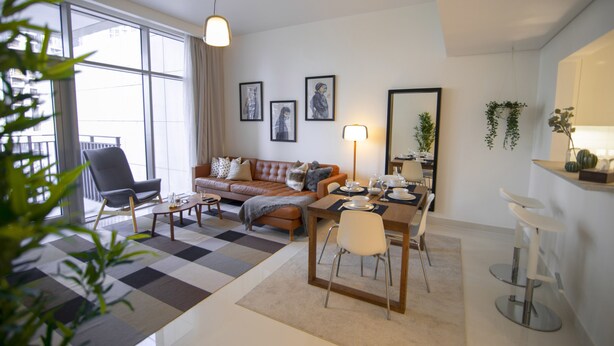 Een moderne woonkamer met een bank, salontafel en eettafel. Er is een groot raam met gordijnen, een plant en ingelijste foto's aan de muur.