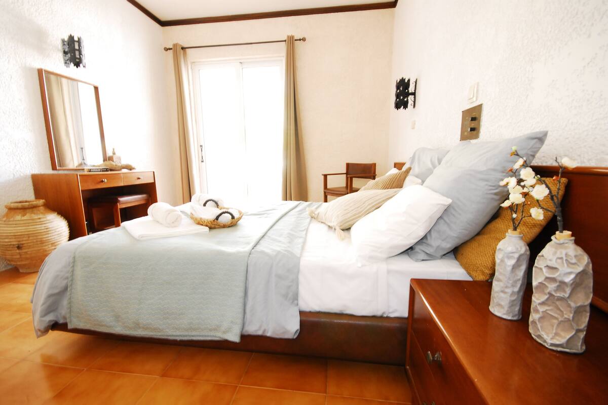 Une chambre à coucher bien rangée, avec un lit, une commode en bois, une chaise et une grande fenêtre avec des rideaux.