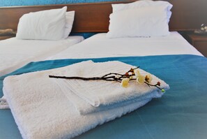 A neatly made bed with white linens and a blue accent, a white towel with a branch and flowers, and a wooden headboard. - Ladikos Garden (Rhodes)