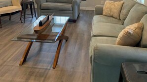 Una sala de estar con una viga de madera en el techo, dos ventanas con persianas, un sofá gris, un sofá beige y una mesa de centro de vidrio.