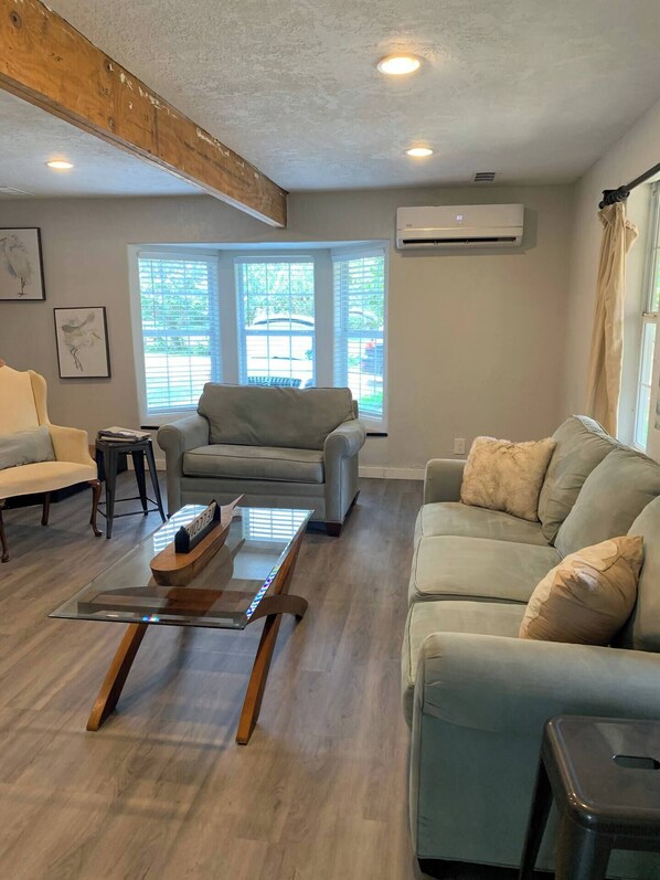 A living room with a wooden ceiling beam, two windows with blinds, a grey sofa, a beige sofa, and a glass coffee table.