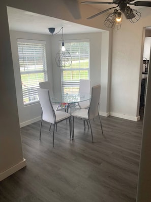 A dining area with a glass table and four chairs, a ceiling fan, and large windows with blinds.