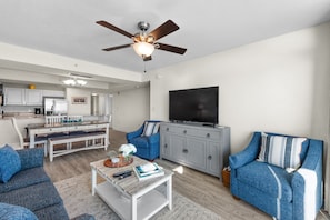 A modern living room with a sofa, coffee table, and TV. There is a dining area with a bench and chairs. - Grande Atlantic 807 - 4 Bedroom Oceanfront Unit (Myrtle Beach)