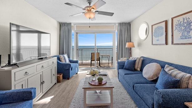 A modern living room with a television, blue sofas, a coffee table, and a balcony with a view of the ocean. - Grande Atlantic 807 - 4 Bedroom Oceanfront Unit (Myrtle Beach)