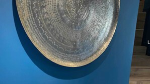 Ein großes, rundes, strukturiertes Metallobjekt an einer blauen Wand, im Vordergrund eine grob bearbeitete Steinskulptur.