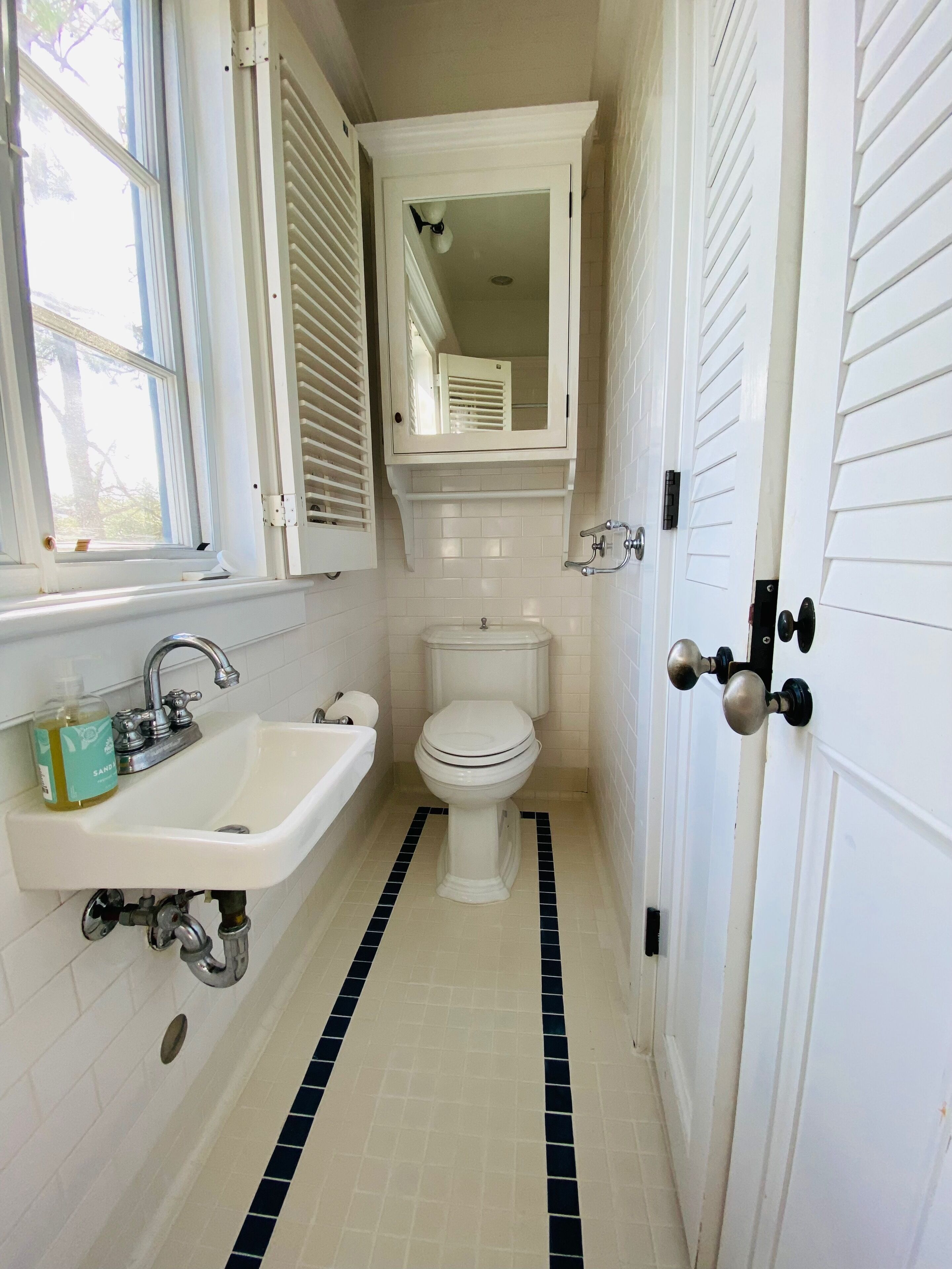 A narrow bathroom with a white sink, toilet, and window with blinds.