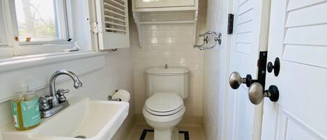 A narrow bathroom with a white sink, toilet, and window with blinds.