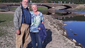 Un hombre y una mujer de pie sobre una costa rocosa, con un puente al fondo.