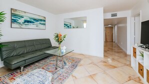 A modern living room with a green sofa, a glass coffee table, and a colorful rug. There is a painting of a seascape on the wall, a mirror, and a hallway leading to another room. - Amazing 2BR APT @Brickell, Free Parking (Miami)