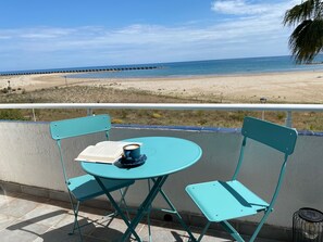 Une table et des chaises en métal turquoise sont placées sur un balcon qui donne sur une plage, avec une jetée en bois visible au loin.