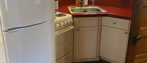 A compact kitchen with a white refrigerator, stove, and sink. There are wooden cabinets and a window with curtains.