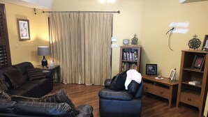 A living room with a black leather sofa, a smaller black leather chair, a wooden side table with a lamp, a bookshelf, and a wooden cabinet with a picture frame.