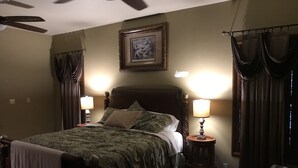 A bedroom with a bed, two bedside lamps, a ceiling fan, a framed picture on the wall, and curtains.