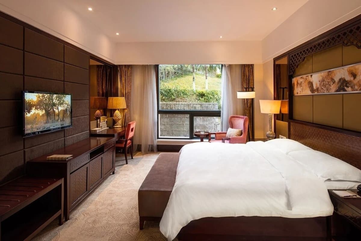 A hotel room with a large bed, a TV mounted on the wall, a desk with a chair, and a window offering a view of greenery.
