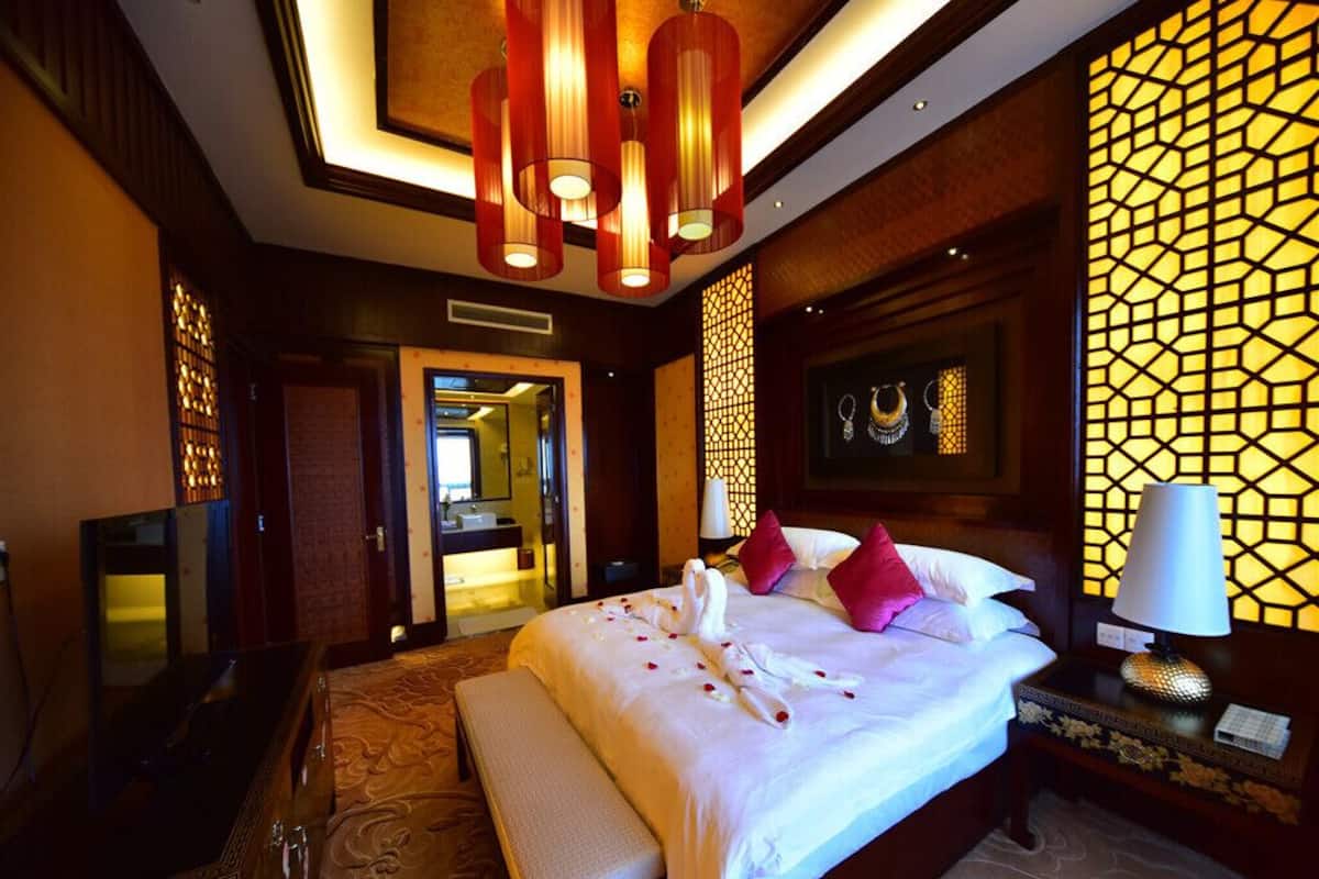 A hotel room with a large bed, a bedside table with a lamp, a TV, and a decorative wall with a geometric pattern.