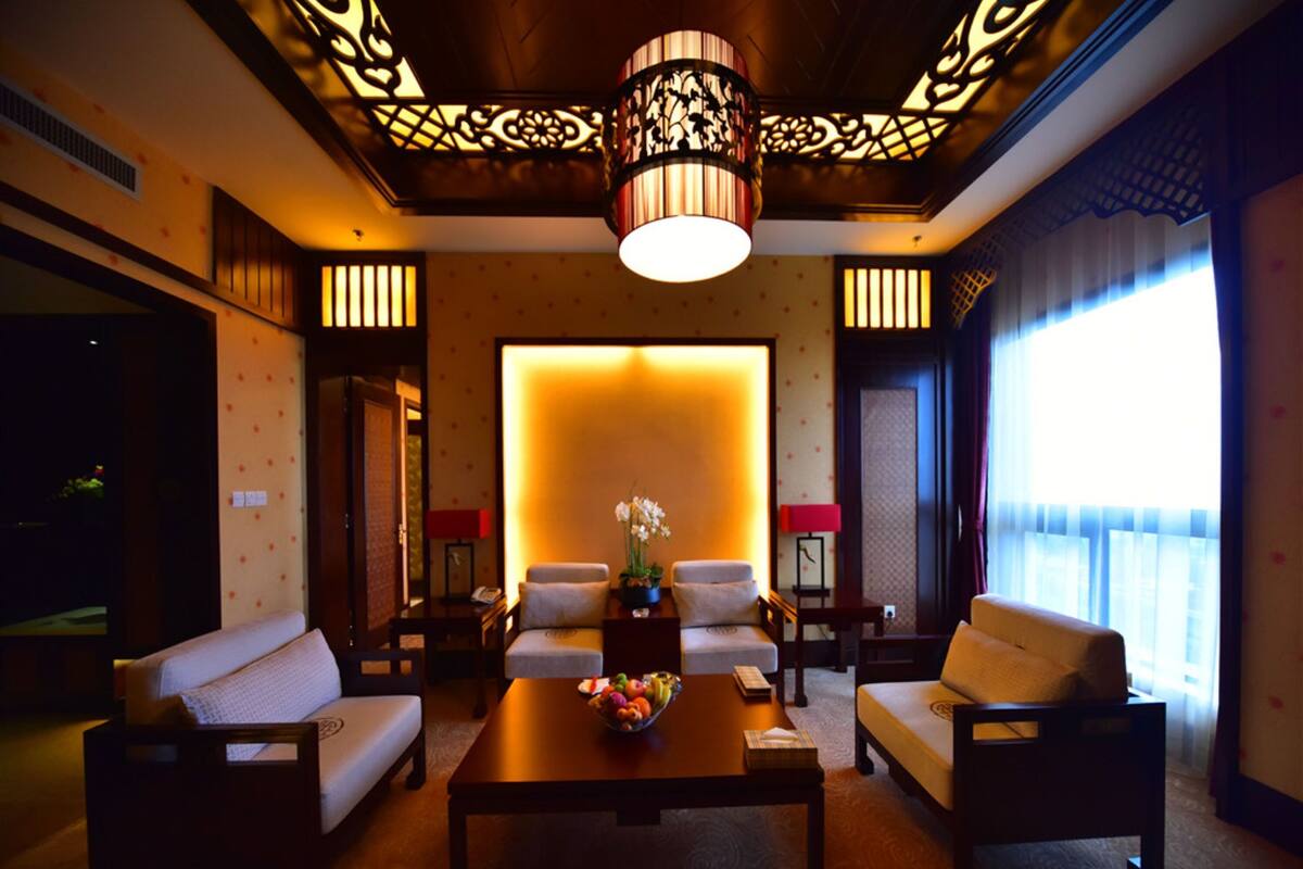 A modern hotel room with a sofa, armchairs, a coffee table, and a decorative ceiling light.