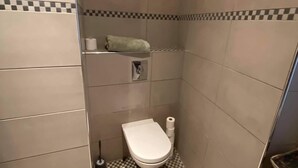Een kleine betegelde badkamer met een zwart-wit geruite vloer, een toilet, een plankje met een handdoek en een toiletrolhouder.