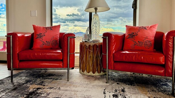 Quarto com duas poltronas de couro vermelho, uma mesa lateral de madeira com abajur e uma grande janela com vista para montanhas e céu nublado.