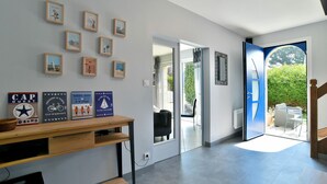 A hallway with a wooden table displaying framed pictures, a blue door leading outside, and a view of a patio area. - Les Daules (Cancale)