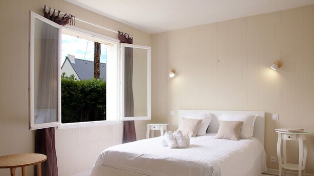 A bedroom with a large bed, two bedside tables, a window with curtains, and a view of greenery outside. - Les Daules (Cancale)
