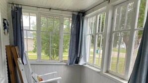 Ein Zimmer mit weißen Wänden, einem grauen Teppich und einem Schaukelstuhl. Es gibt Fenster mit Vorhängen und ein an die Wand gelehntes Holzstuhls.