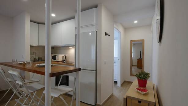 Une cuisine moderne avec des armoires blanches, un plan de travail en bois et un réfrigérateur. On y trouve un coin repas avec des chaises blanches et une petite table. Une plante en pot est posée sur une table d’appoint.