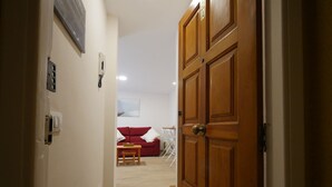 Un couloir mène à un espace de vie comprenant un canapé rouge, une petite table et une chaise. Une porte en bois se trouve à droite et un thermostat fixé au mur, à gauche.