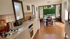 A modern kitchen with a central island, a dining area with a table and chairs, and a view of a garden through an open door. - Soustons Grande Propriété Dans un Écrin de Verdure en Lisière de Forêt (Soustons)