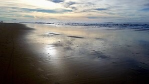 Een strand met nat zand en voetafdrukken, een bewolkte lucht en verafgelegen bergen.