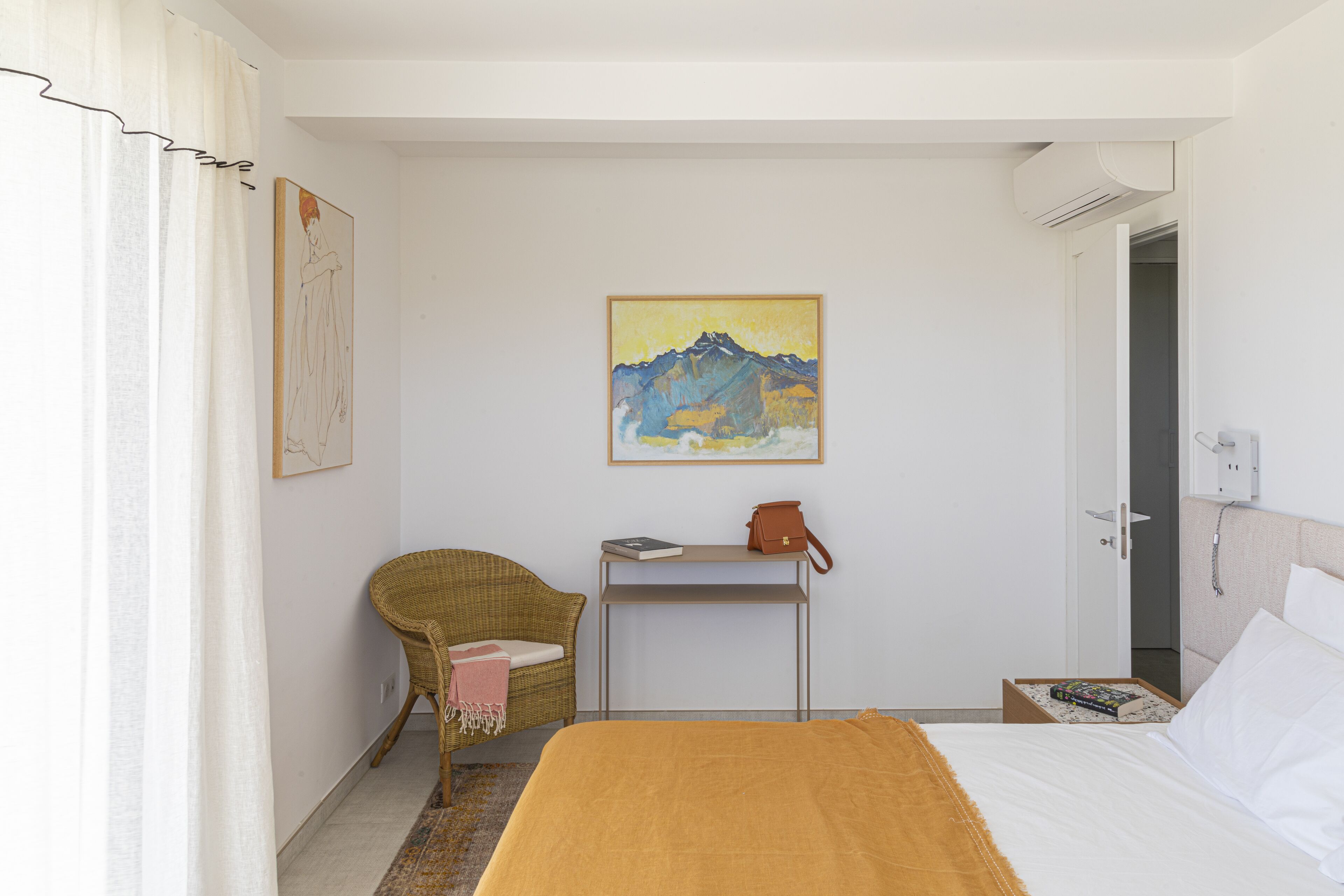 Une chambre d’hôtel avec un lit, une chaise, une table avec un livre et un sac, un tableau au mur et une porte.