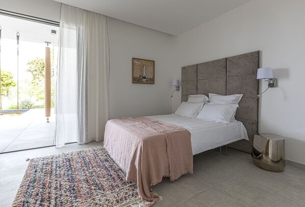 A modern bedroom with a large bed, a headboard, a bedside lamp, and a view of the outdoors. - Casa Baia Estate ★★★★★ (Calvi)