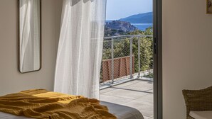 A bedroom with a bed, a window with sheer curtains, and a view of a coastal town. - Casa Baia Estate ★★★★★ (Calvi)