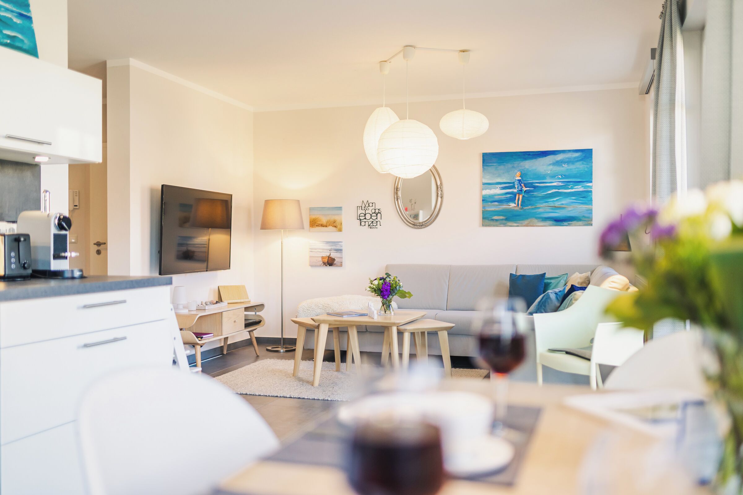 A modern living room with a sofa, coffee table, and a dining area with chairs. There is a painting on the wall and a television mounted above the sofa.