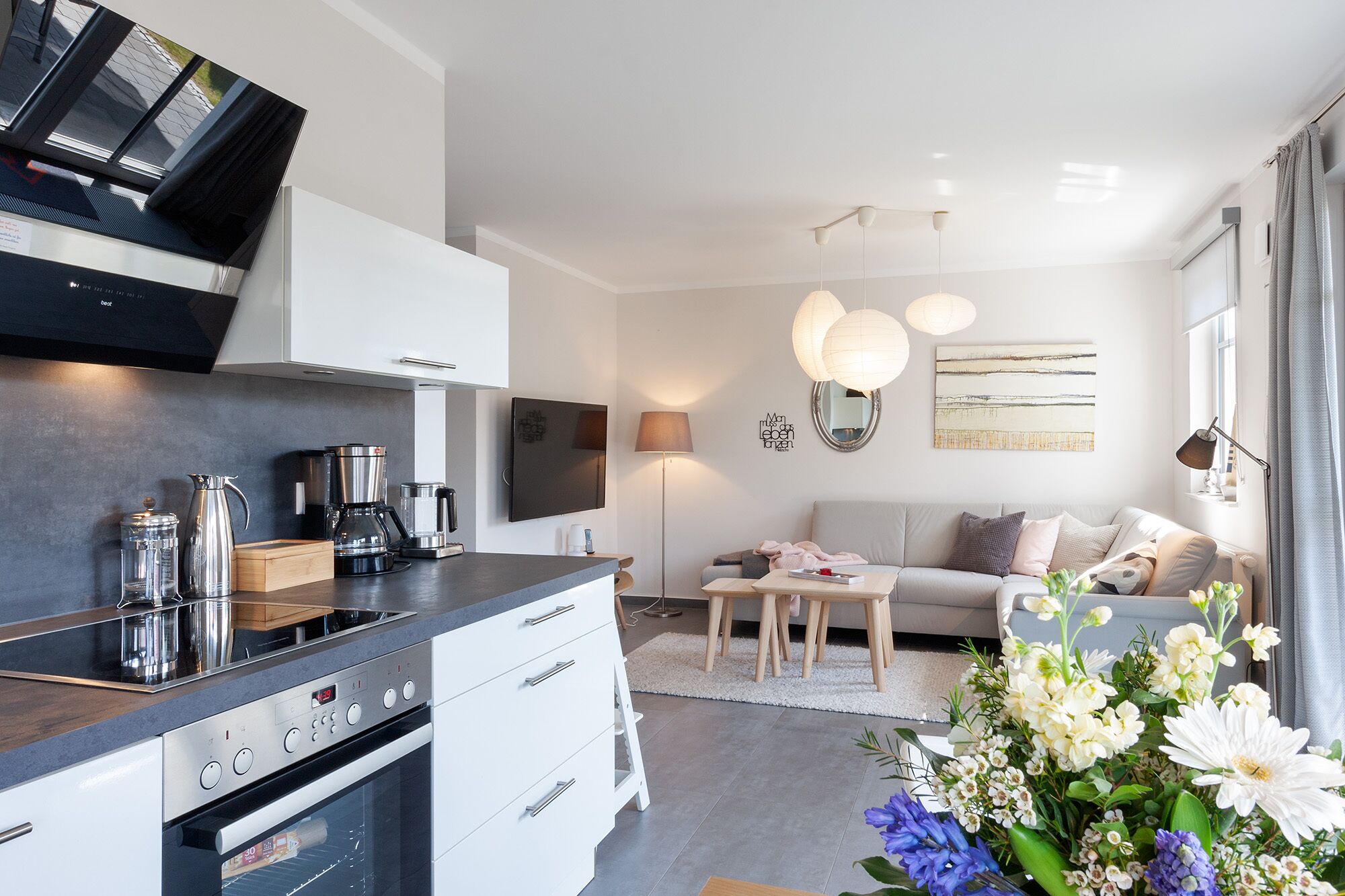A modern kitchen with a built-in oven and a coffee maker. There is a sofa and a dining table in the living area, and a large window with curtains.