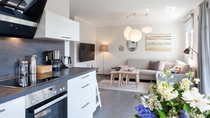 A modern kitchen with a built-in oven and a coffee maker. There is a sofa and a dining table in the living area, and a large window with curtains. - Fewo am Horizont No3 Exklusivgenießen in Zingst (Zingst)
