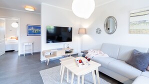 A modern living room with a flat-screen TV, a sofa, a coffee table, and a dining area with a lamp and a small table. - Fewo am Horizont No3 Exklusivgenießen in Zingst (Zingst)